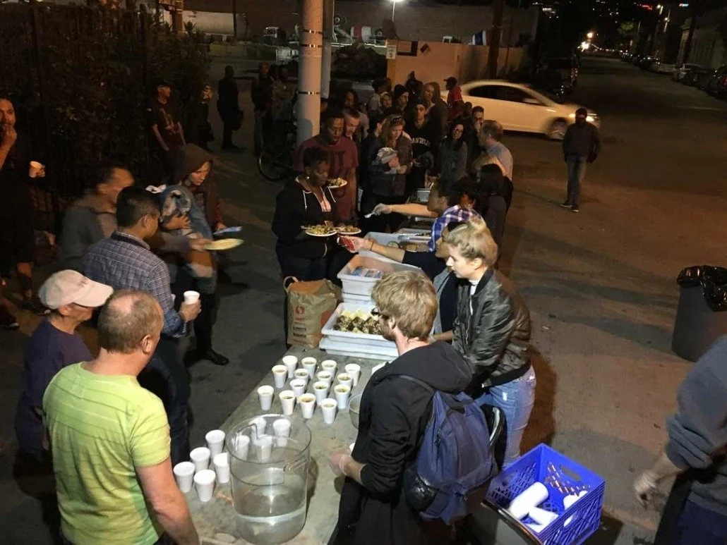 People standing in a line outdoors at night, receiving food and drinks from volunteers at a community service event.