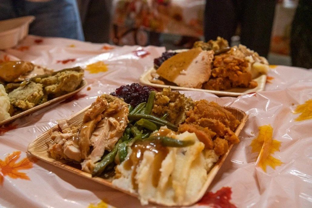 Plates of Thanksgiving dinner with turkey, stuffing, mashed potatoes, green beans, and cranberry sauce on a table covered with autumn-themed tablecloth.
