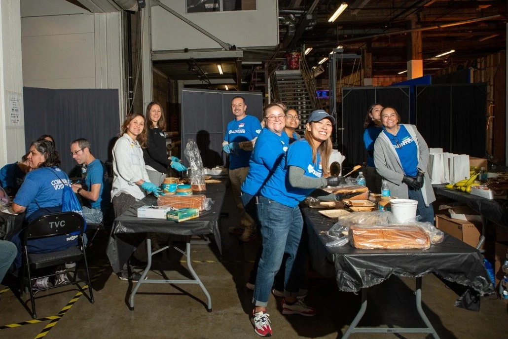 Group of volunteers preparing food at a community event in an indoor space, some wearing blue volunteer shirts and aprons, with tables filled with bread and supplies.