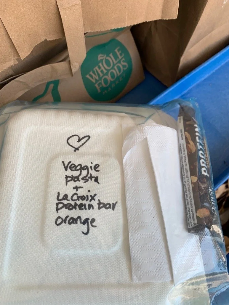 A white plastic container with handwritten notes indicating it contains veggie pasta and La Croix protein bar, placed inside a clear resealable bag. In the background, Whole Foods Market paper bag and other packages are visible.