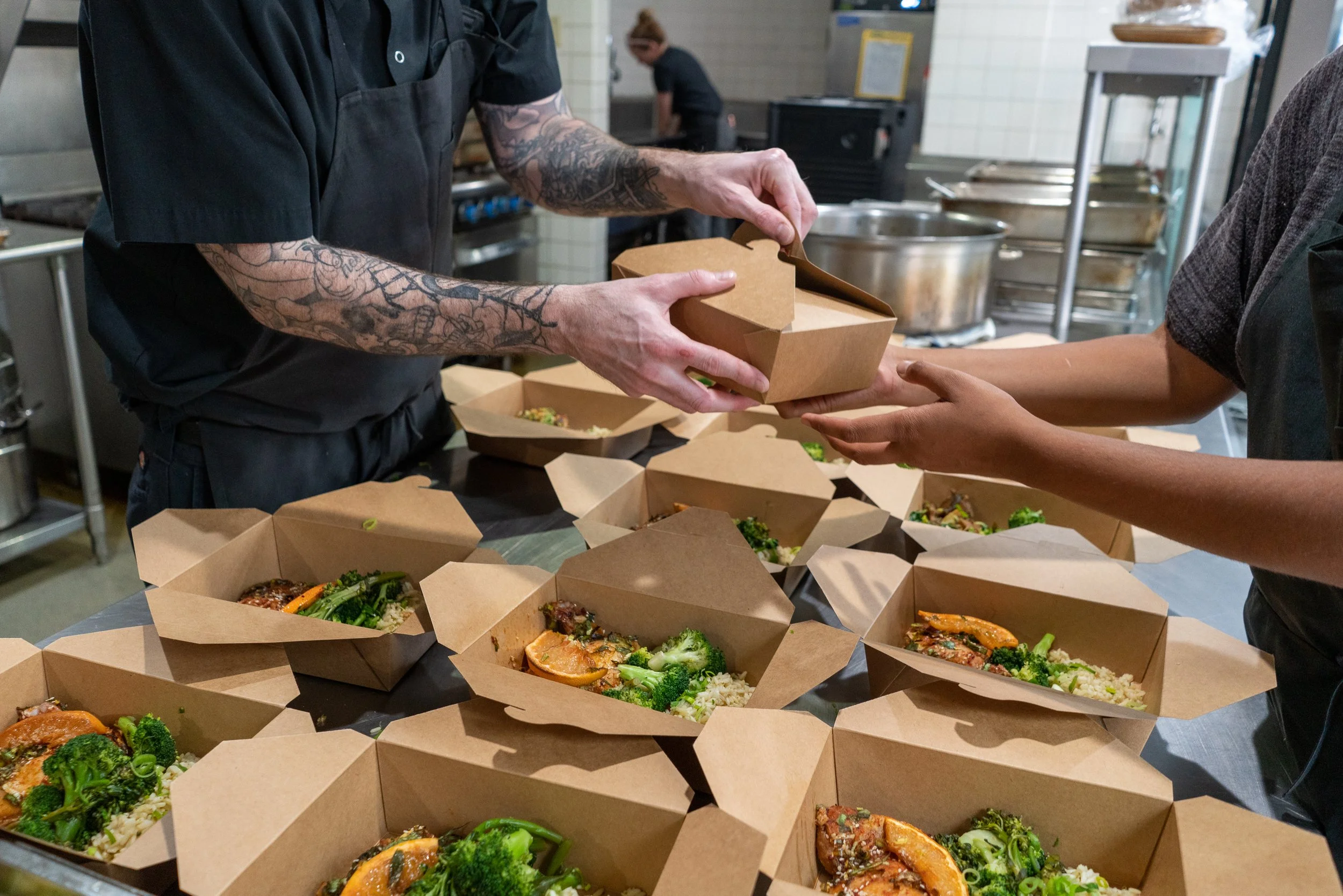Person packaging food in eco-friendly brown boxes with green vegetables and rice on a stainless steel countertop in a kitchen.
