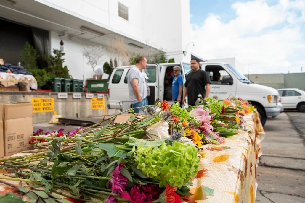 Table covered with colorful flowers in front of a parking lot where people are gathered near a white van, with boxes and signs in the background.
