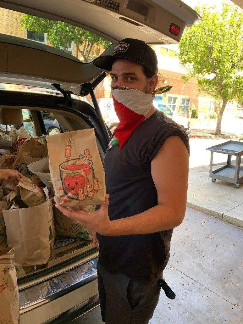A young man wearing a black t-shirt, a black cap, and a cloth face covering stands next to an open car trunk filled with paper bags. He is holding a paper bag with a drawing of a smiling cup of tomato juice with a smiling tomato and small carrots. Th