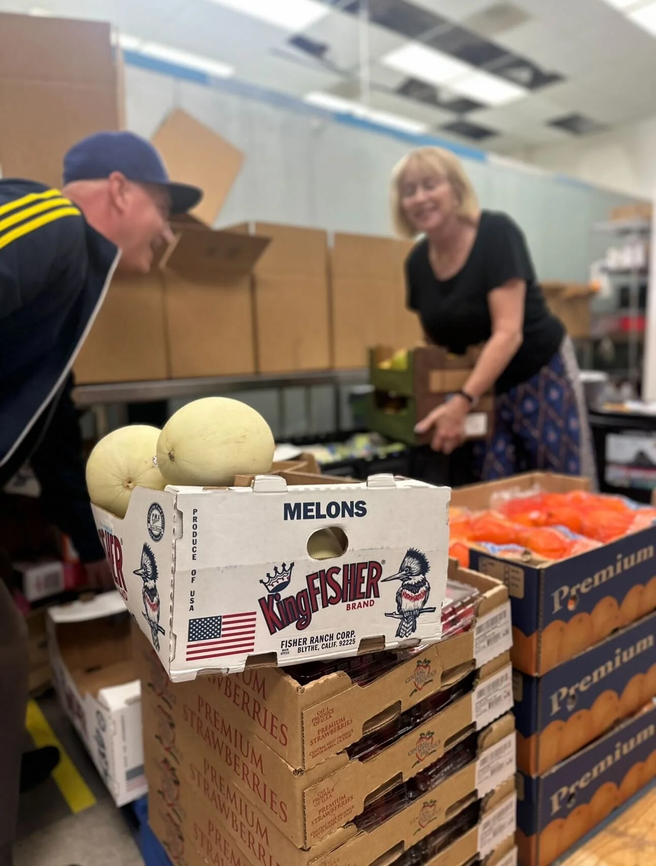 A grocery store scene with a cardboard box of melons, and two women in the background, one smiling and the other leaning over the boxes.