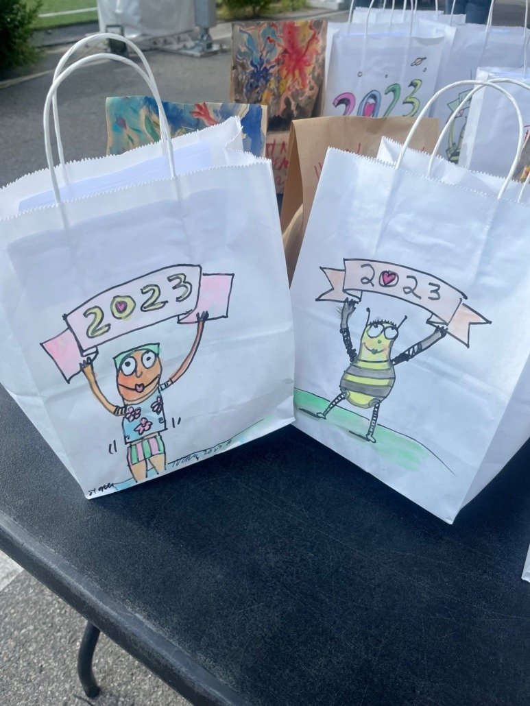 Two white paper bags with colorful cartoon drawings of children holding a banner that reads '2023.' One child is wearing a hat and shorts, and the other is in a striped outfit. Additional bags with similar artwork are in the background.