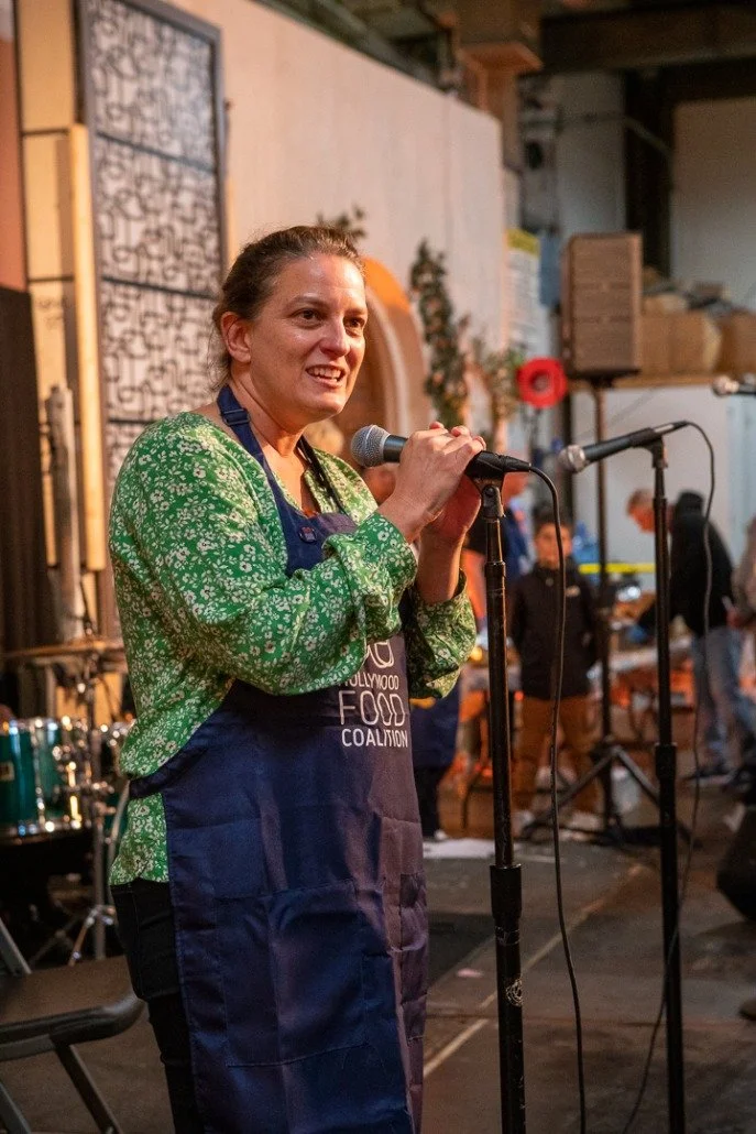 A woman in a green floral shirt and blue apron speaking into a microphone at an indoor event.