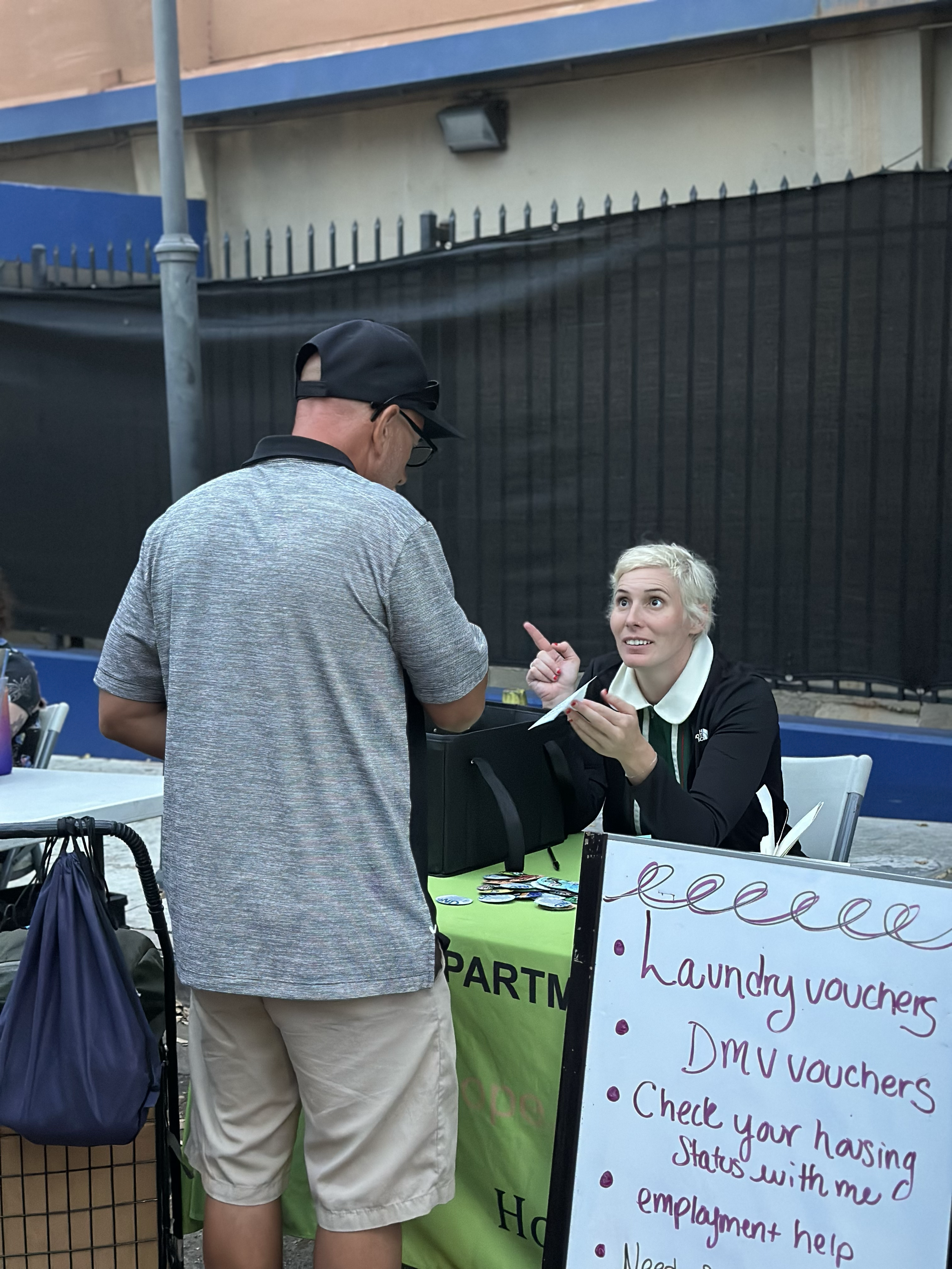 A woman with short blonde hair engaging with a man at an outdoor table with a sign offering laundry and DMV vouchers, and employment help.