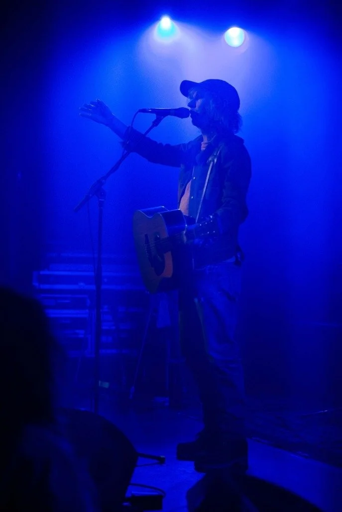 A person performing on stage with a guitar under blue lights, wearing a baseball cap and casual clothing.