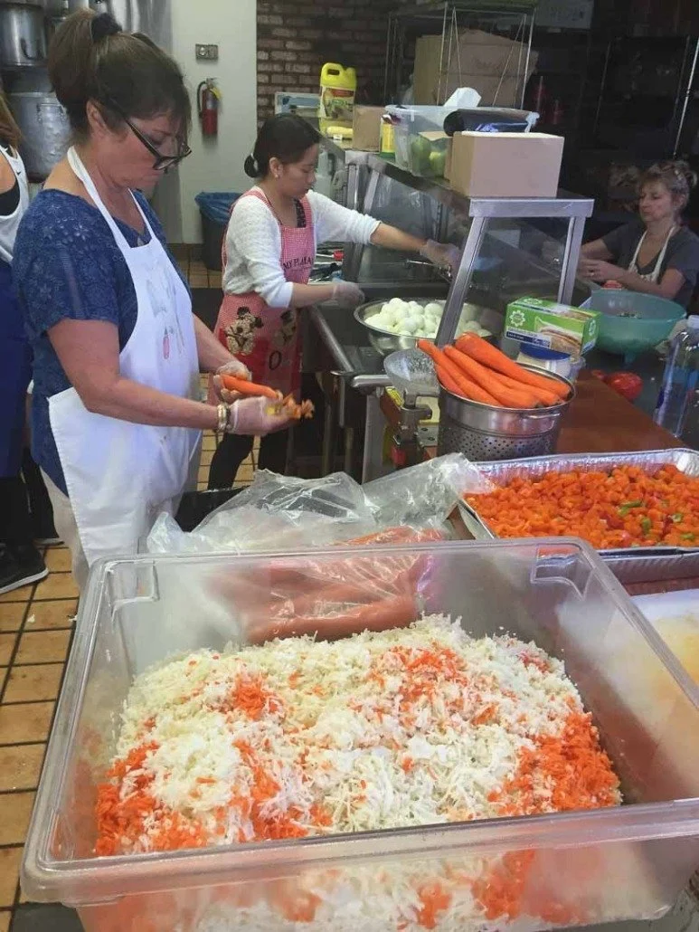 Three women preparing food in a kitchen or cafeteria, with vegetables and shredded cheese on a counter in front of them.