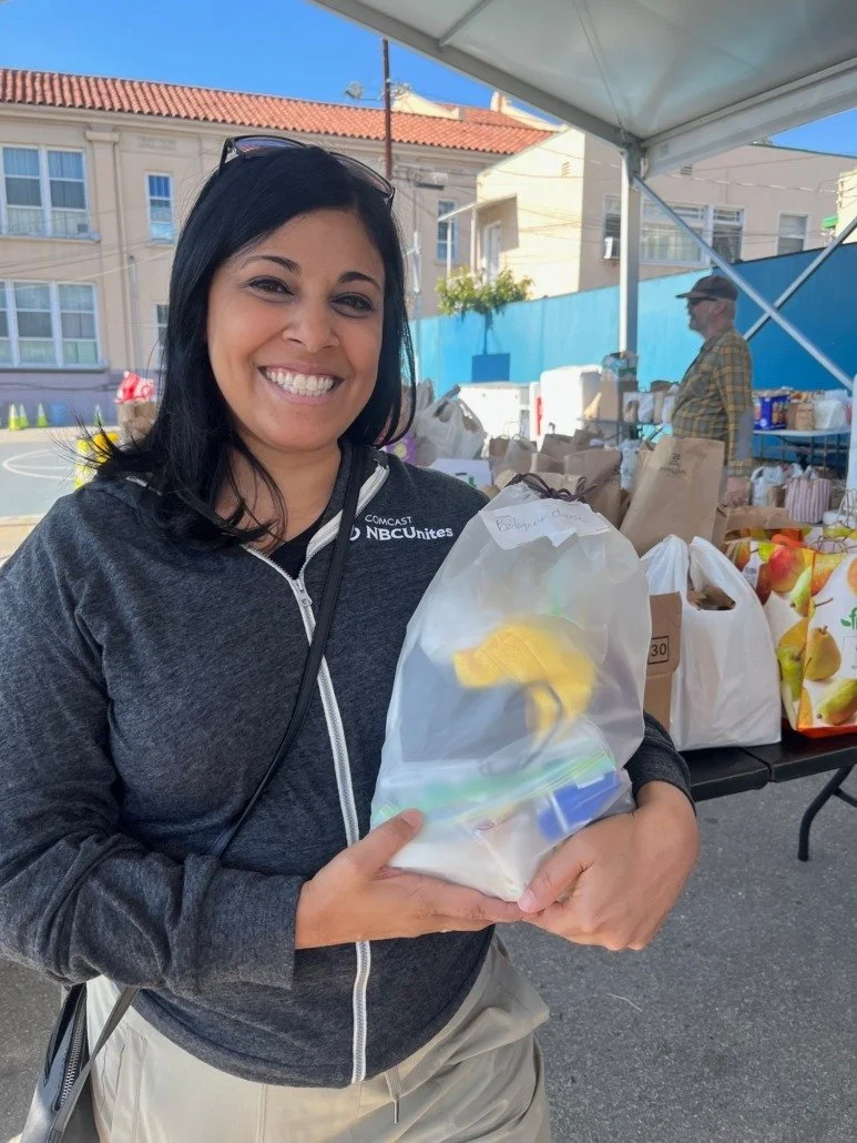 A smiling woman with black hair and sunglasses on her head holding a plastic bag with groceries at an outdoor market. She is wearing a dark zip-up jacket with the NBCUniversal logo.