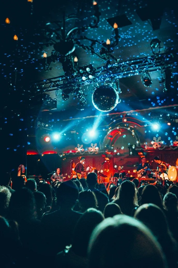 A concert stage decorated with Christmas-themed decorations such as snowflakes and holiday lights. Musicians are performing with instruments, and a large crowd is watching. The lighting is vibrant, with blue and red lights and a large disco ball hang