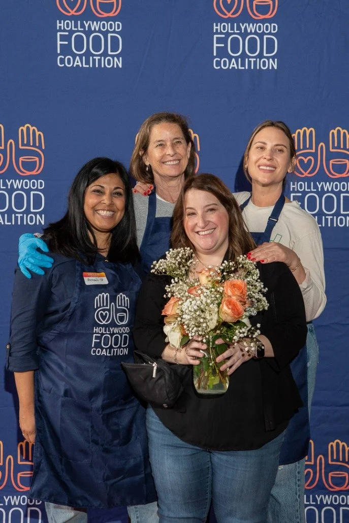 Four women smiling and posing for a photo at a Hollywood Food Coalition event. One woman in the front holds a bouquet of peach roses and white flowers. They stand in front of a blue backdrop with the organization’s logo.