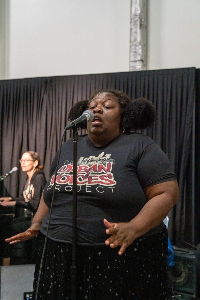 A woman singing passionately into a microphone, wearing a black t-shirt with the words 'The Brazilian Voices Project' on it, standing in front of a black curtain, with another person playing the piano in the background.