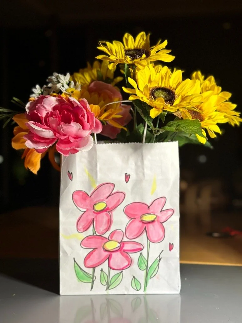 A paper bag with a colorful floral drawing holds a bouquet of real flowers including pink peonies, yellow sunflowers, and white daisies, set against a dark background.