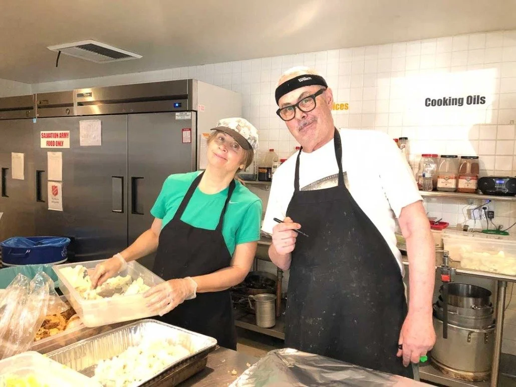 Two people in a kitchen preparing food, one woman and one man, both wearing aprons and hats.
