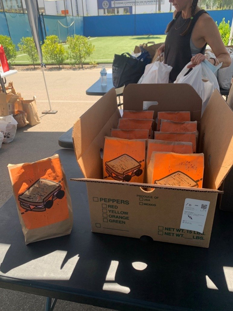 Box of paper bags with illustrated sandwiches on them, placed on a table at an outdoor event.
