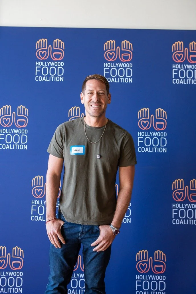 A man smiling in front of a blue backdrop with Hollywood Food Coalition logo, wearing a green t-shirt, jeans, and a wristwatch.