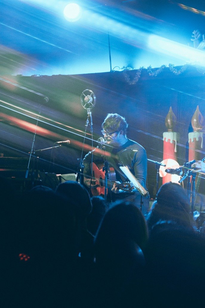 A musician performing on stage with a guitar and microphone, illuminated by colorful stage lights, with audience members visible in silhouette and holiday-themed decorations in the background.