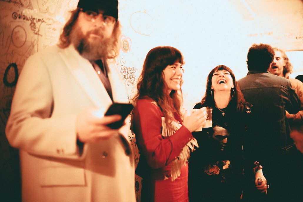 Group of people socializing in a dimly lit room with graffiti on the wall, some holding drinks, smiling and laughing.