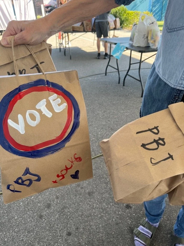 Person handing over paper bags labeled 'PBJ' and a paper bag decorated with a red and blue circle with 'VOTE' written inside at an outdoor event.