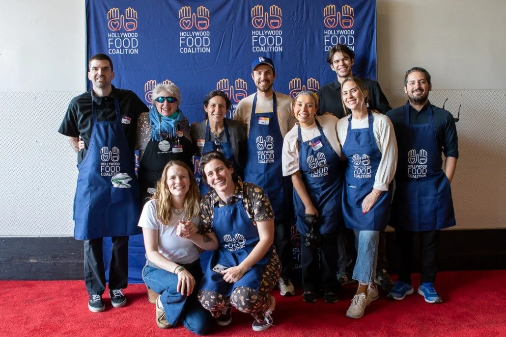 Group of people at a Hollywood Food Coalition event, some wearing aprons, standing in front of a blue backdrop with the coalition's logo, smiling and posing for the photo.