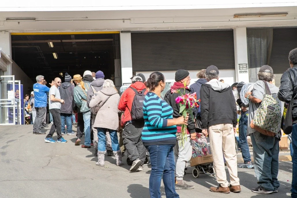People standing in line outside a large building with an open roller shutter door.
