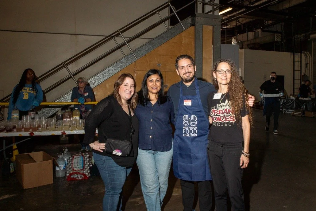 Four people smiling and posing for a photo in an industrial space, with a table of drinks and a staircase in the background. One person is wearing an apron with "Hollywood Food Coalition" on it, and another is in a t-shirt with "Urban Voices Project"