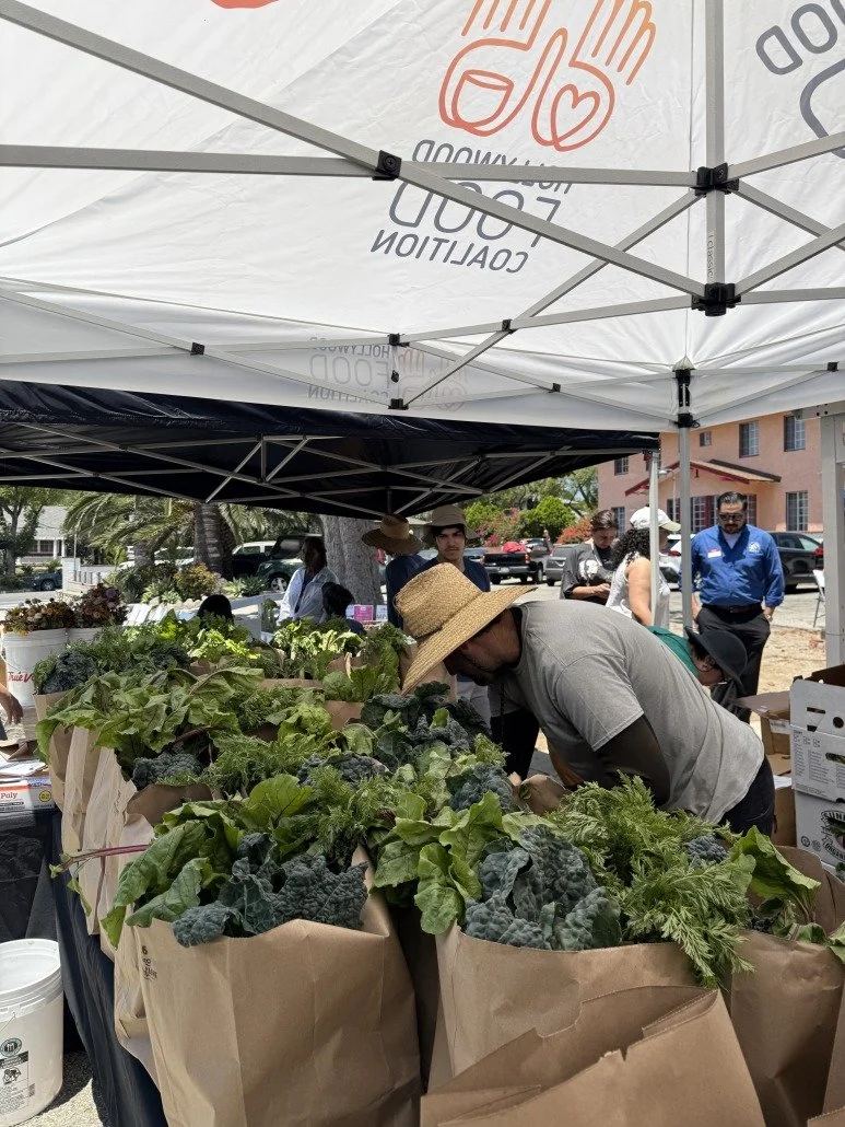 People shopping for fresh vegetables and greens at an outdoor market stall under a white canopy tent.
