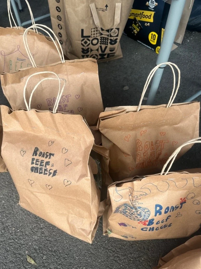 Multiple brown paper shopping bags with handles, labeled "Roast Beef & Cheese" and decorated with small hearts, placed on a rough asphalt surface.