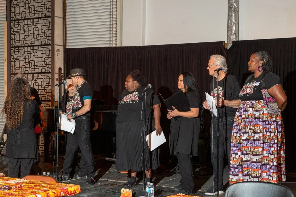 Group of people on stage participating in an event, some holding papers, with a woman at a microphone and others standing in line, against a backdrop of black curtains.