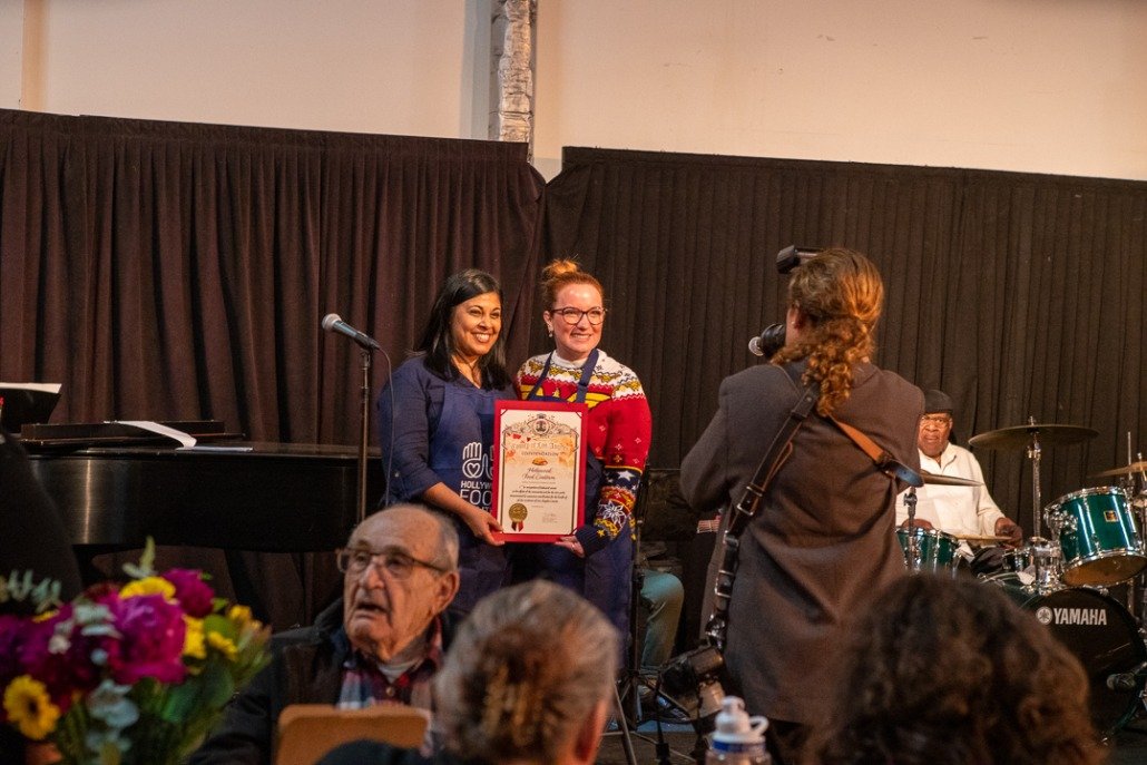 Two women are standing on stage, smiling, with one holding a framed certificate. A photographer is taking their picture. There is a man playing drums in the background, and other people seated in the audience. The stage has dark curtains.