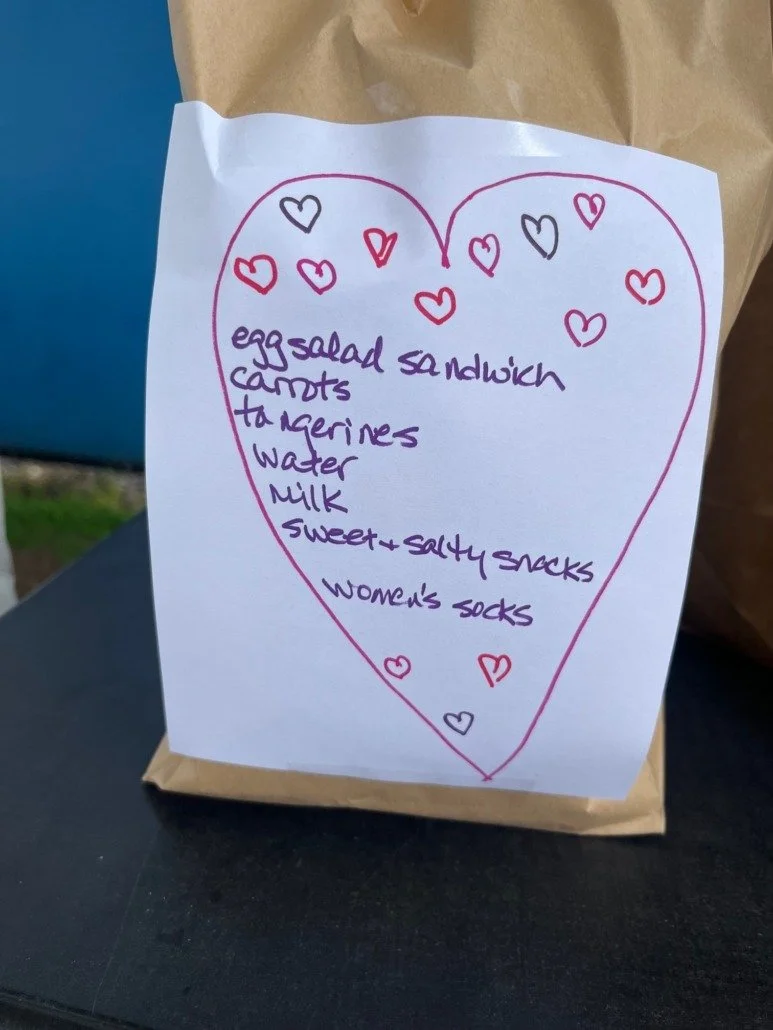 A handwritten list of grocery items on a piece of paper attached to a paper bag, surrounded by a pink heart outline with small hearts around it.