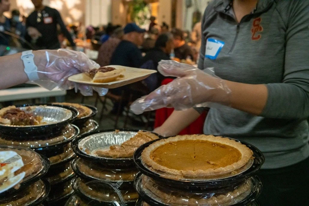 Food distribution at a community event, with a person wearing gloves handing out slices of pie or casserole on a paper plate over stacked pies or casseroles.