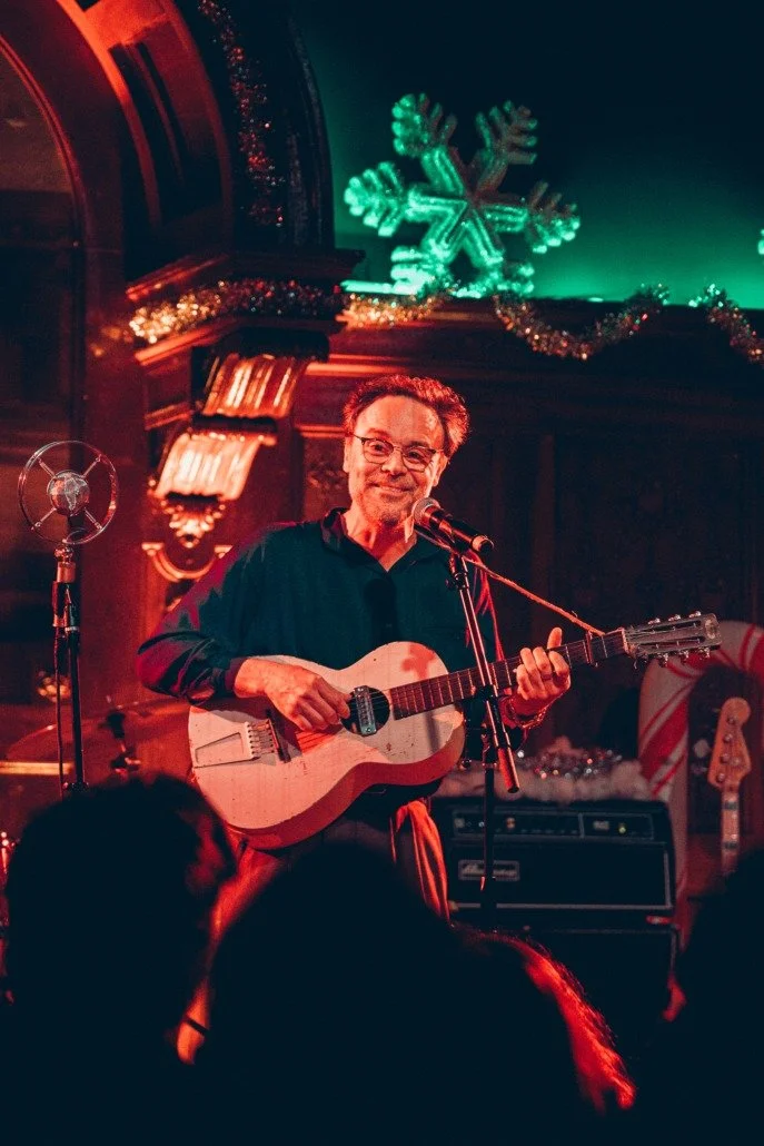 A man with glasses smiling and playing an acoustic guitar on stage, with a microphone in front of him, during a festive event decorated with Christmas ornaments, including a large glowing snowflake and tinsel.