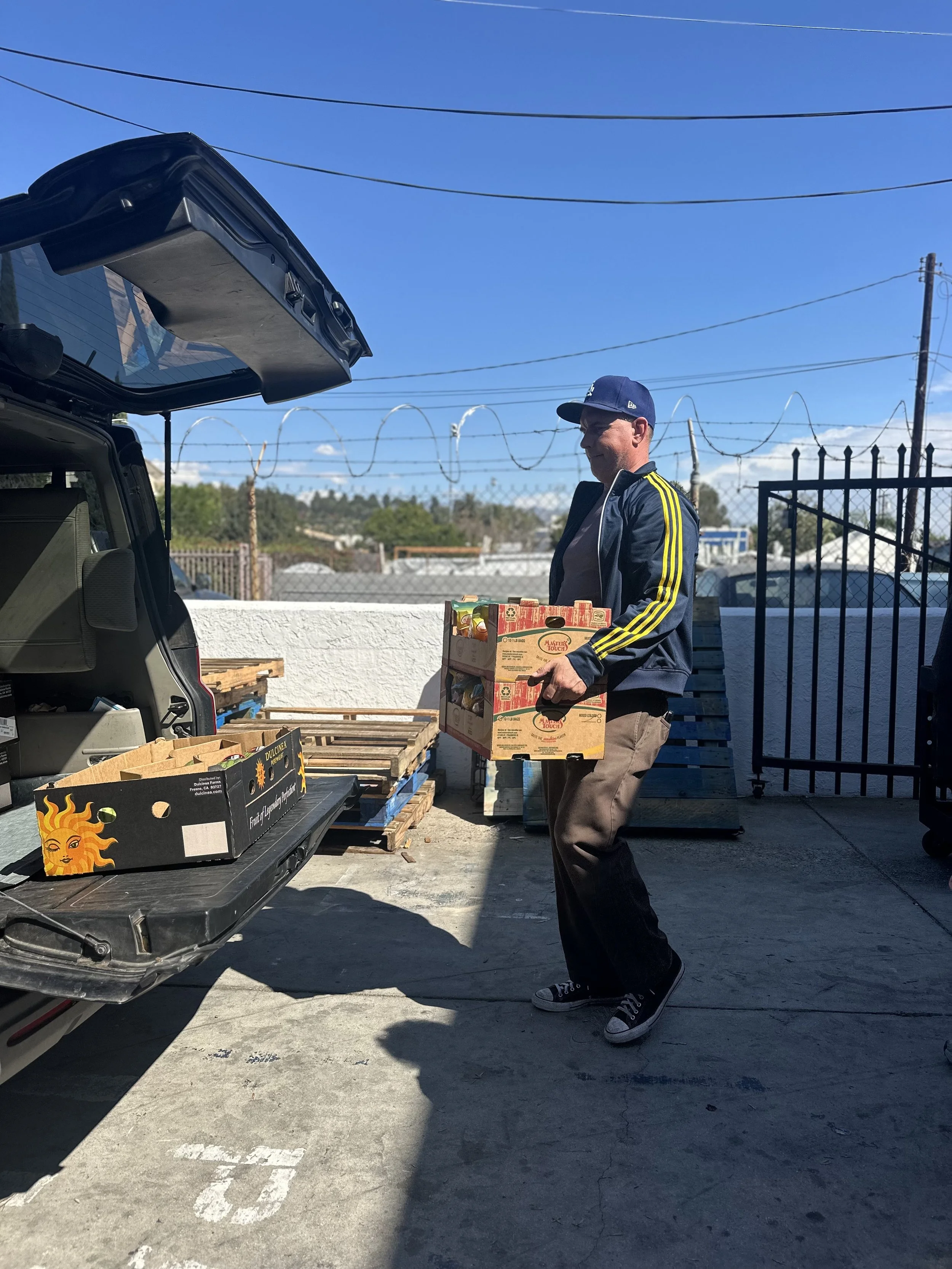 A man wearing a blue cap, black jacket with yellow stripes, and brown pants is carrying a box of produce while standing next to an open vehicle trunk in an outdoor area with a gate, barbed wire, and pallets.