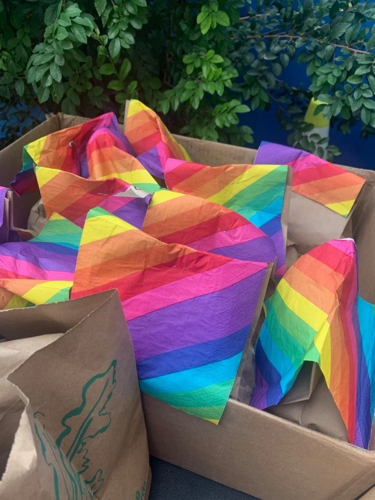 Colorful rainbow-themed paper wrappers inside a cardboard box with green foliage in the background.