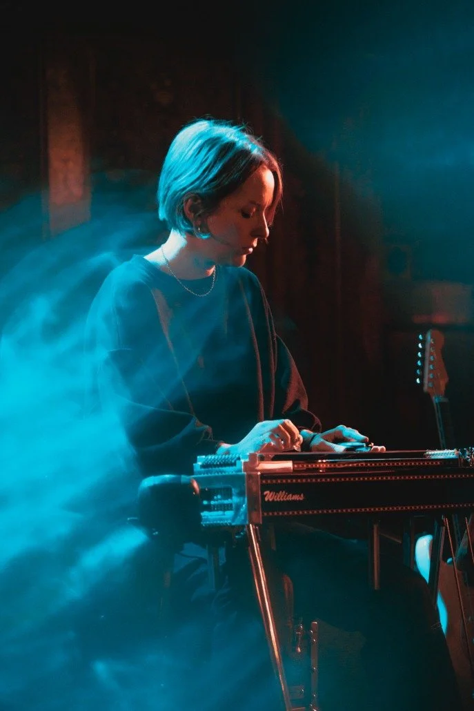 A woman with short hair playing a steel guitar on stage with blue lighting and smoke effects.