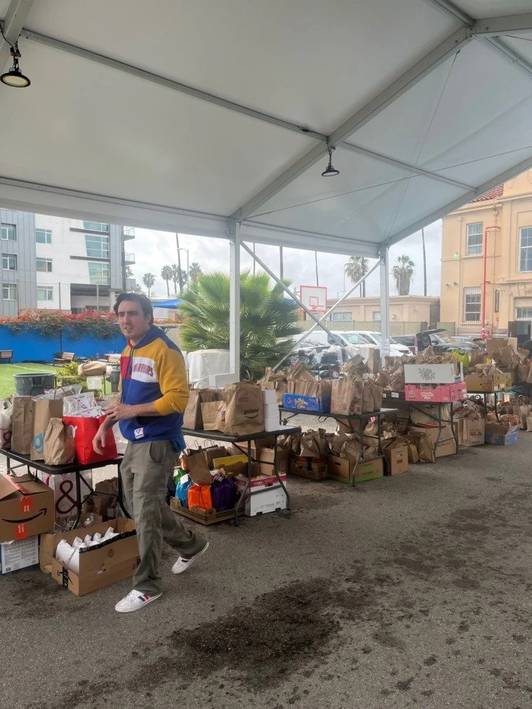 A young man standing under a large outdoor canopy, surrounded by tables filled with paper bags, boxes, and supplies, with a background of buildings, palm trees, and parked cars.