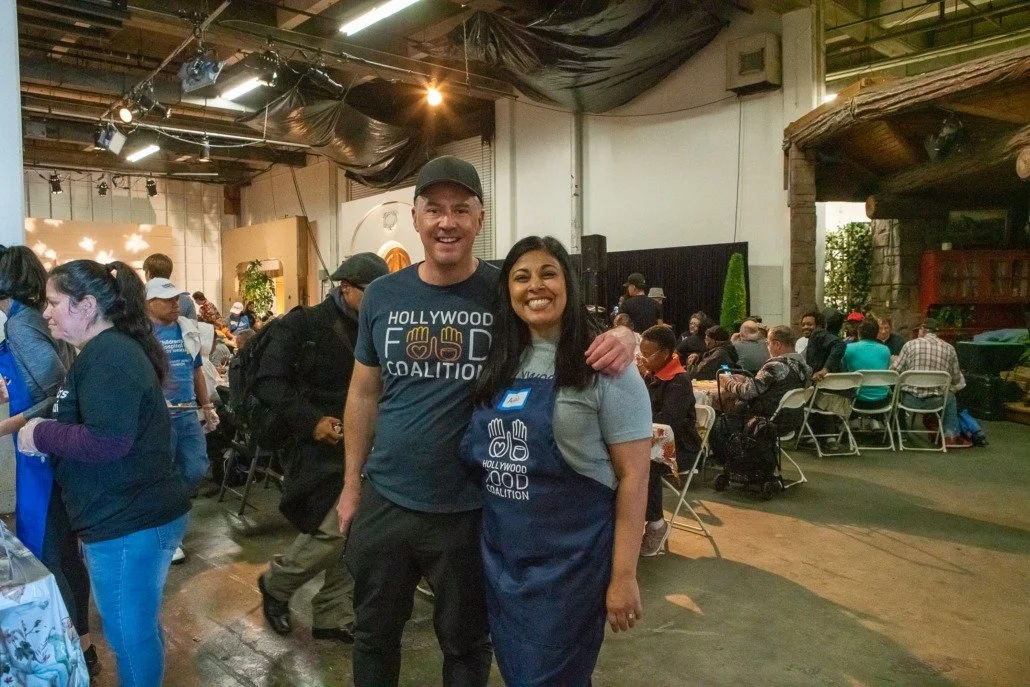 Two people standing together smiling in a crowded indoor event space, with many people seated at tables in the background.