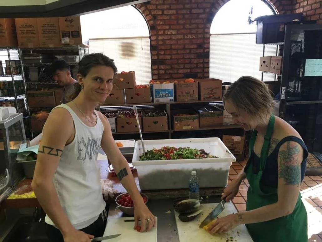 Two women preparing food in a kitchen, one chopping vegetables and the other smiling at the camera, with a large container of salad and other food items visible behind them.