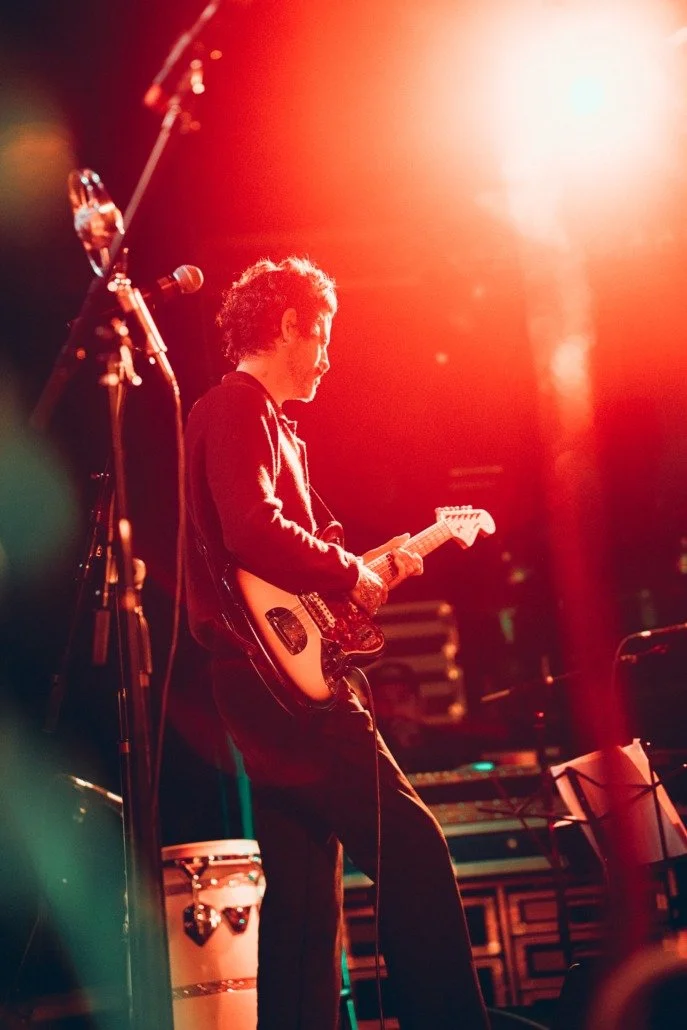 A musician playing an electric guitar on stage under warm orange stage lighting.