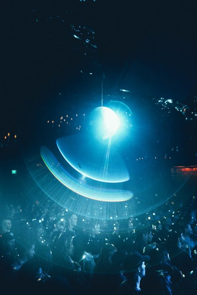 A large disco ball hanging from the ceiling of a dark club or concert venue, reflecting bright blue and white light across the room filled with people.
