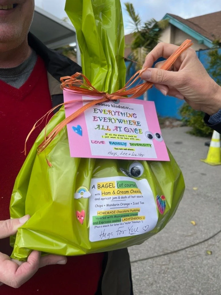 Person holding a gift bag decorated with colorful stickers and handwritten notes, with a card attached that has a message about kindness, love, family, and diversity, and a menu for bagel, ham and cream cheese, chips, mandarins, hot sauce, homemade p