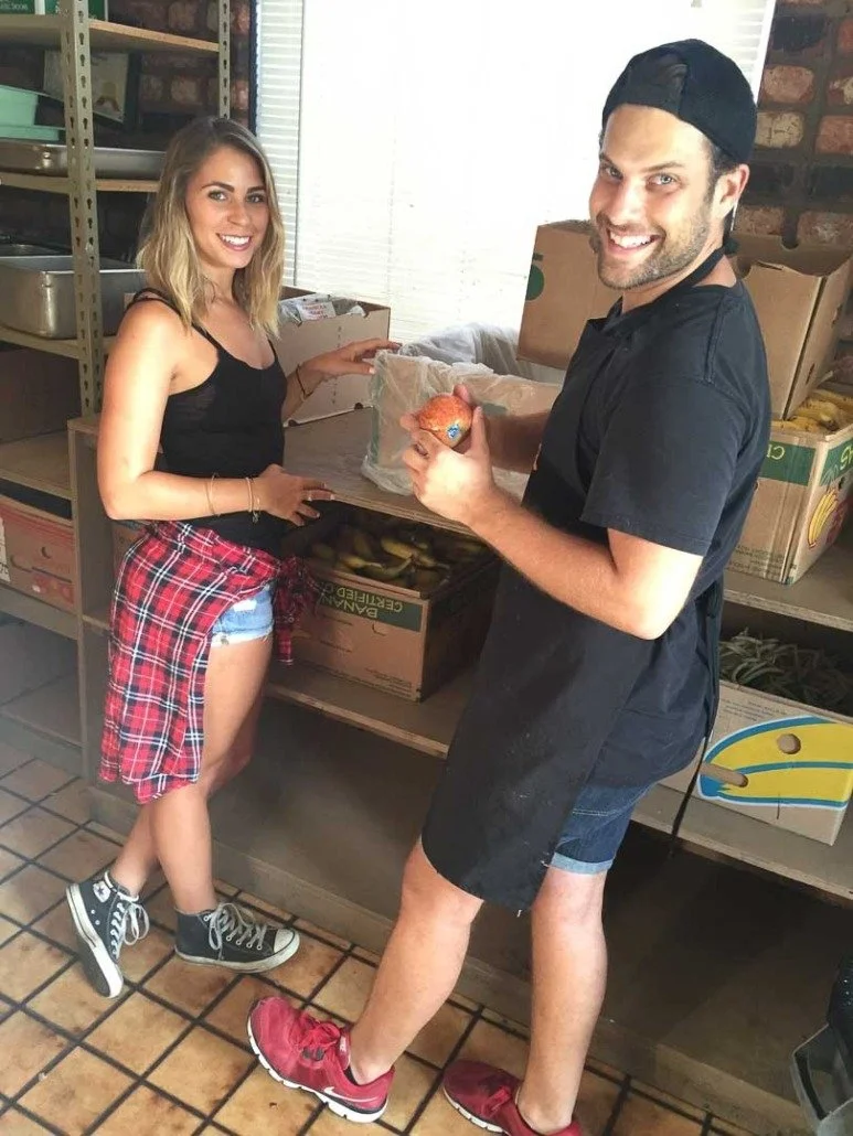 A man and a woman standing in a storage room near shelves filled with boxes and produce. The woman is smiling, wearing a black tank top, denim shorts, a red plaid shirt tied around her waist, and Converse sneakers. The man is holding a bag of onions,