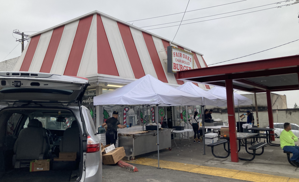 Outdoor food stand with white tents and people working, in front of a red and white striped building with a sign advertising burgers at Fair Oaks.