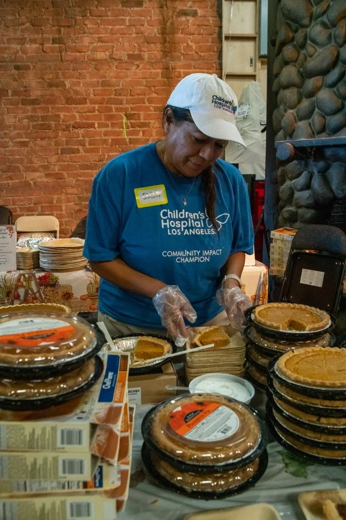 A woman wearing a Children's Hospital Los Angeles t-shirt and cap, and clear gloves, preparing slices of pumpkin pie at a community event with stacked pies and disposable plates on the table.