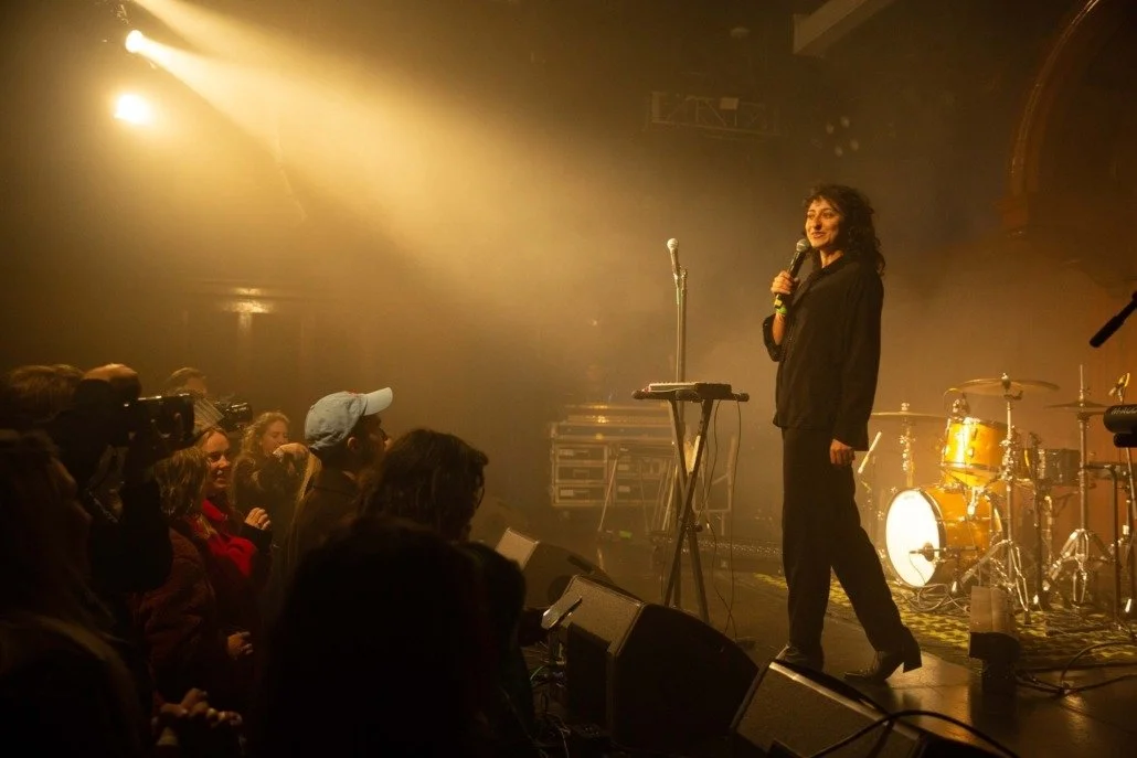 A female performer standing on stage with a microphone, keyboard, and drum set in the background, facing an audience in a dimly lit concert venue.