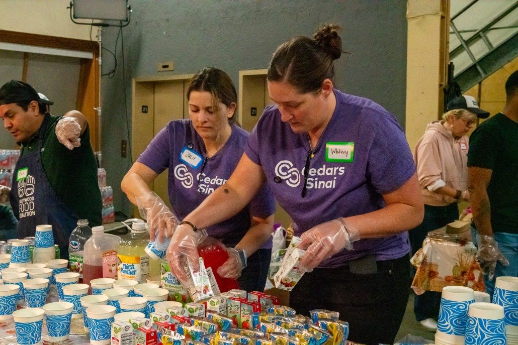 People sorting and preparing food and beverages at a community service event, wearing purple 'Cedar Sinai' shirts and plastic gloves.