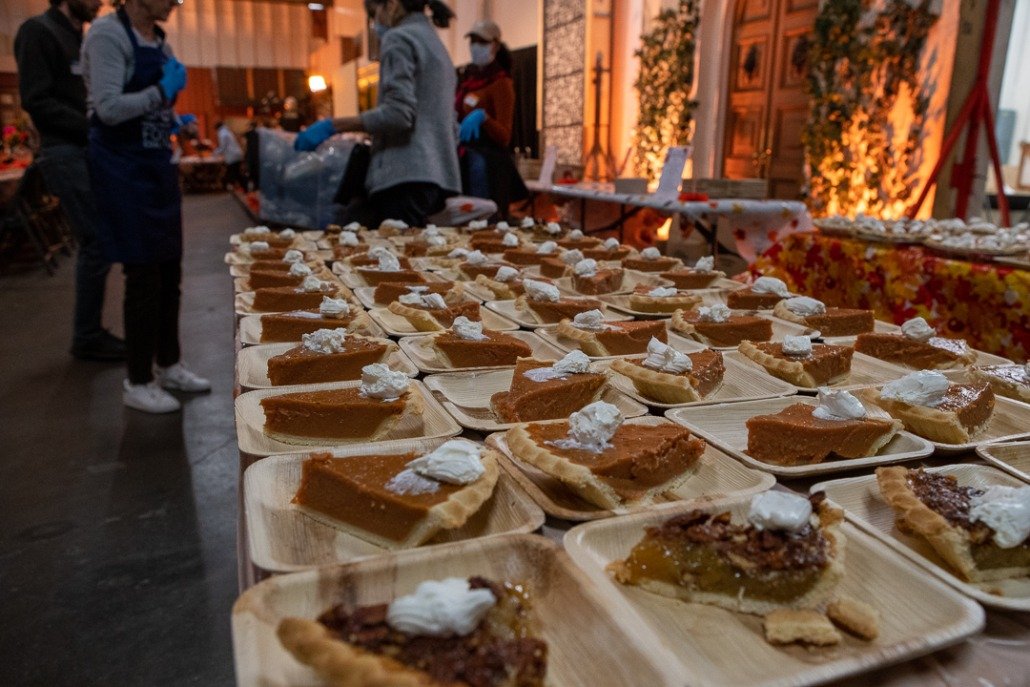 Several slices of pumpkin and pecan pie on wooden trays at a donation or serving table in an indoor setting with people in the background.