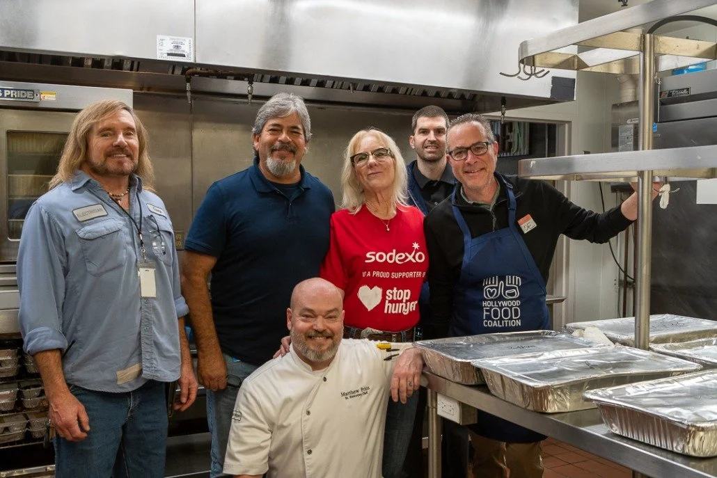 Group of six people in a commercial kitchen, standing around stainless steel counters with trays of prepared food, smiling at the camera.